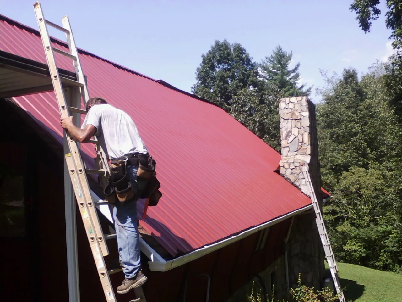 Roofer on a ladder beside a red metal roof and stone chimney — Soffit & Fascia Repair by Roofing Brothers in Chatsworth